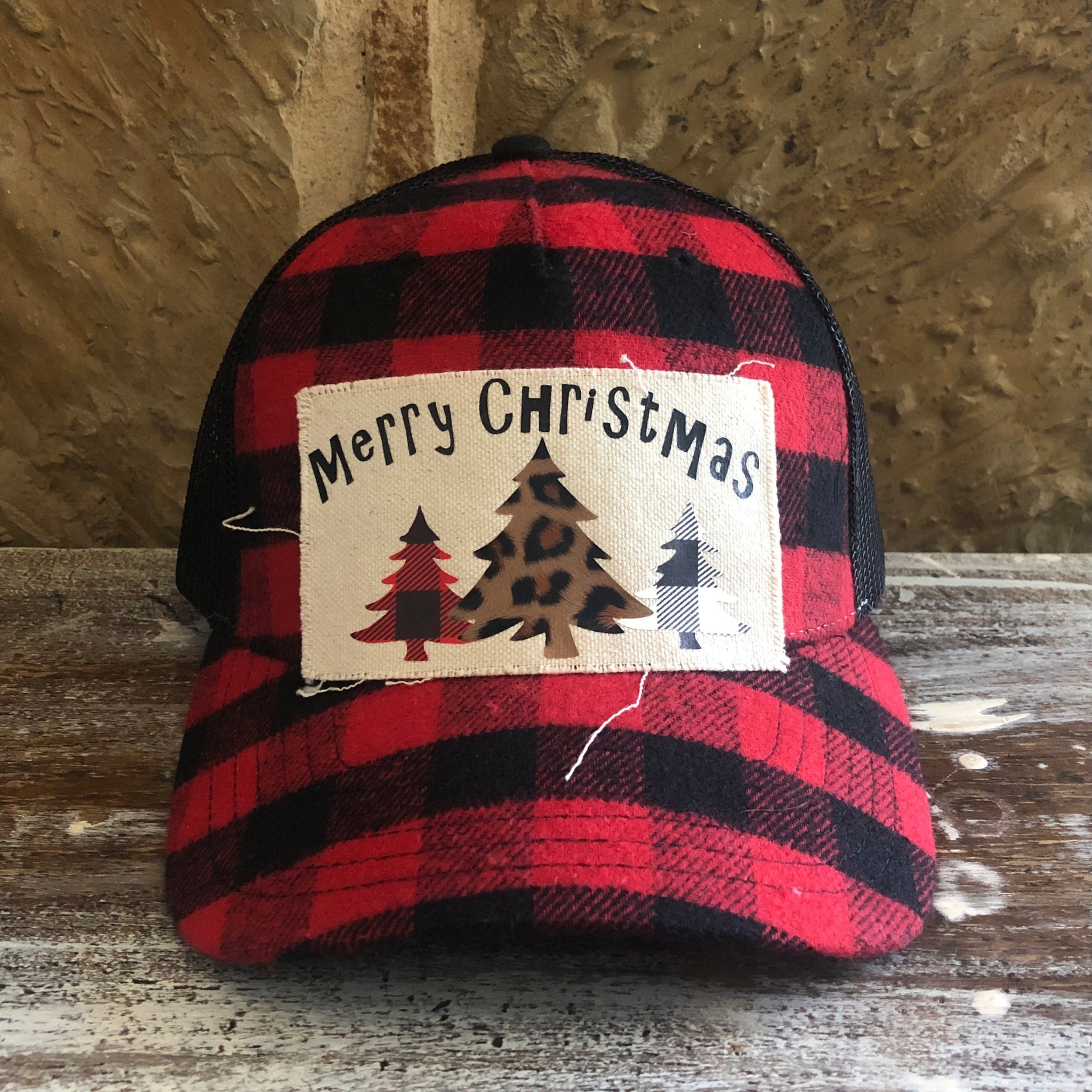 Red and black plaid cap with a 'Merry Christmas' patch featuring trees on a wooden surface.